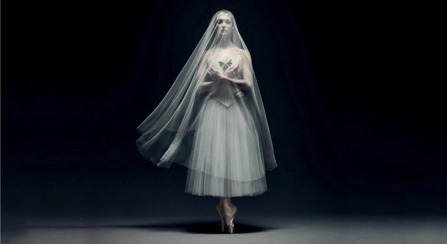 A woman in a white dress stands against a black background with her hands crossed in front of her. A long white veil covers her face and flows in the air. 