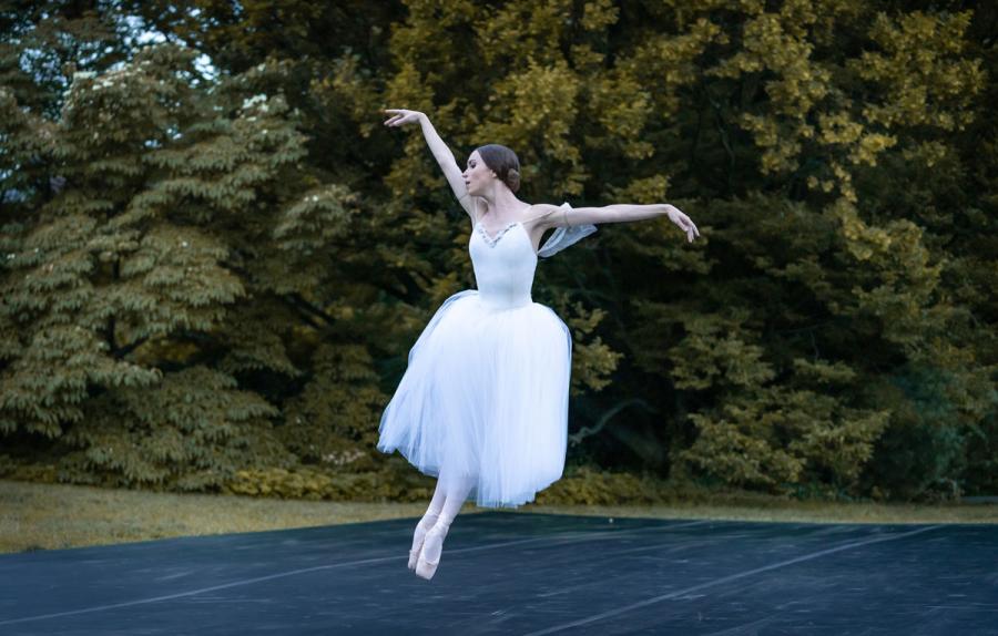 A dancer in a white dress in mid-jump, feet pointed and together to one side, arms open above and to the side.