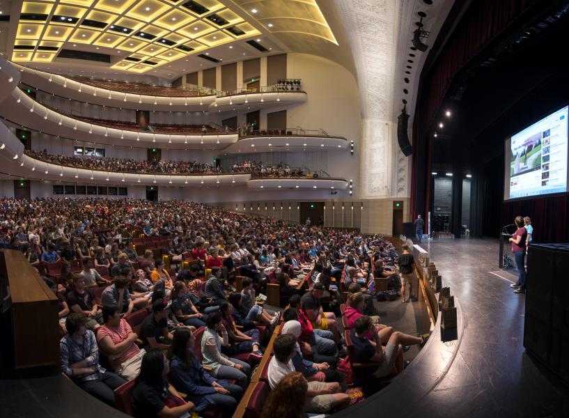 A view of the Carlson Family Stage and audience in the auditorium shot at a very wide angle. The audience is full and two people stand on the stage giving a presentation.