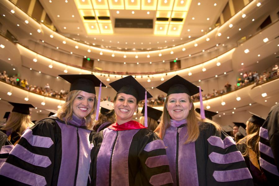 Three light-skinned women wearing black and purple Graduation caps and gowns smile towards the camera. The balconies of the auditorium are lit up behind them and filled with audience members.