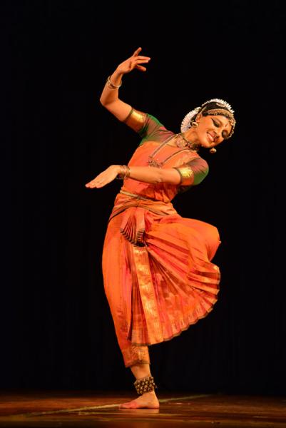 Female dancer in red stands on one foot, the other foot on her knee, leaning to the side with rounded arms in front of her, while she smiles, gazing down over her shoulder. 