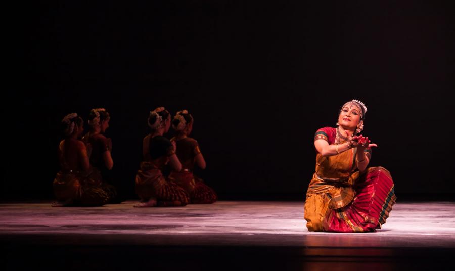 Female dancer in red traditional Indian dress kneels on one knee, hands reaching forward with palms up like a cup