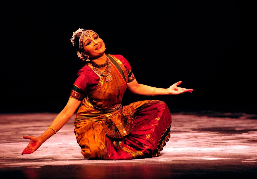 Female dancer in red traditional Indian dress sits cross-legged, smiling and leaning slightly to one side with her elbows in and hands reaching out.