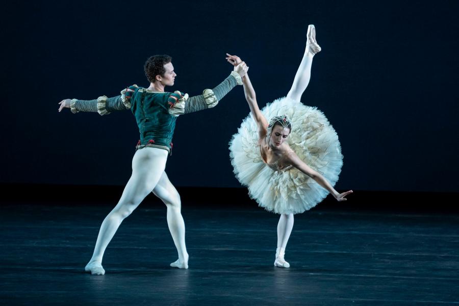 Male dancer in white tights and emerald and gold top coat lunges holding the hand of female dancer in white and gold poses in a high arabesque with her torso parallel to the floor