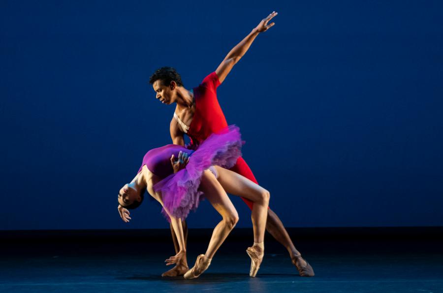 A male dancer wearing a red costume leans with one arm out and holding a female dancer wearing a purple costume with one arm as she bends backwards on pointe.