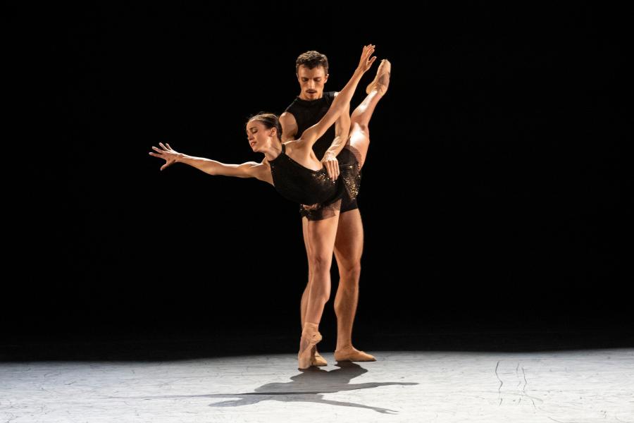 A male dancer holds a female dancer in as she stand on pointe with arms out and leg in a high arabesque.. They are under a spotlight on a dark stage.