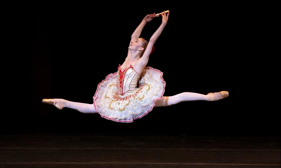 Dancer in white, red and gold leaps with legs straight and arms up above her head