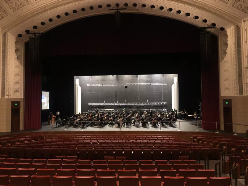 A view of the Carlson Family Stage with rows of chairs and people sitting perpendicular to the empty seats of the auditorium. A video plays on the side of the stage for the audience sitting on stage.