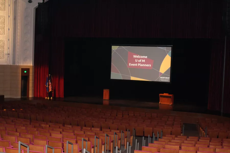 A dark and empty auditorium. On the Carlson Family Stage there is a screen saying "Welcome U of M Event Planners".
