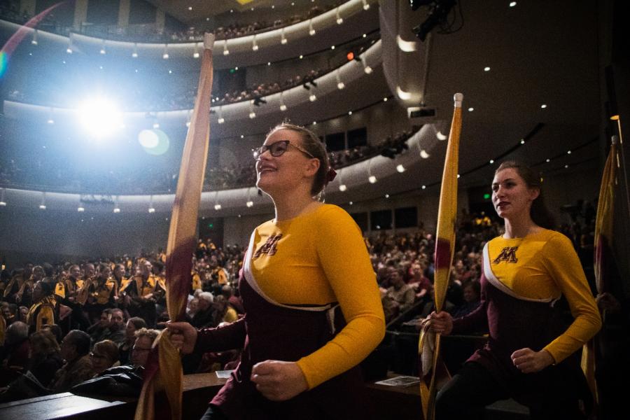 In a full auditorium two female members of the University of Minnesota Marching Band carrying Gold flags smile and march in a line towards the Carlson Family Stage.