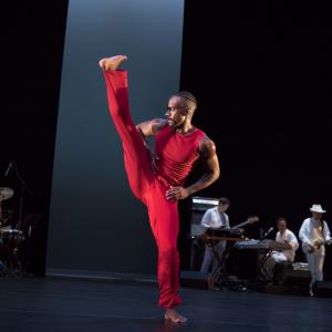 A male dancer wearing all red kicks his leg high on stage as a band plays in the background.
