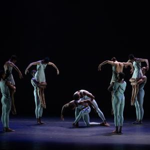 4 female dancers are lifted by their partners, they have their heads down and arms out. They form a circle around another pair in a hug pose at the center.