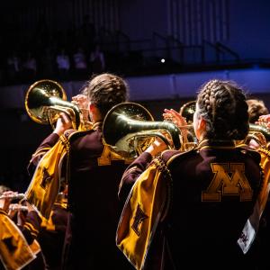 The backs of a three UMN Marching Band members performing on stage, wearing maroon and gold, waving large flags into the air.